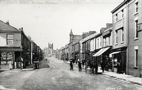 Postcard photograph entitled Church Street, Seaham Harbour 32699 J.V., showing a street running away from the camera towards a church which blocks the far end of the street; shops can be seen on both sides of the road, but only the name of H. Wright, Butcher, can be discerned; indistinguishable figures can be seen on the pavements and a horse and cart is on the right of the picture