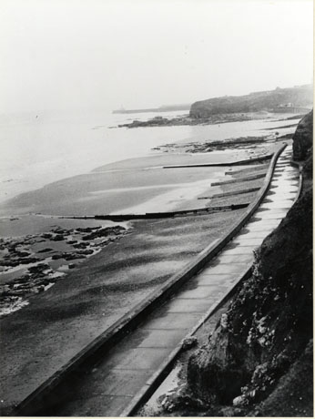 Photograph, taken from above, of a promenade running from bottom left to top right of the photograph, with the sand and the sea on the left and a cliff in the distance on the right; the photograph has been identified as View along the Promenade to Featherbed Rock