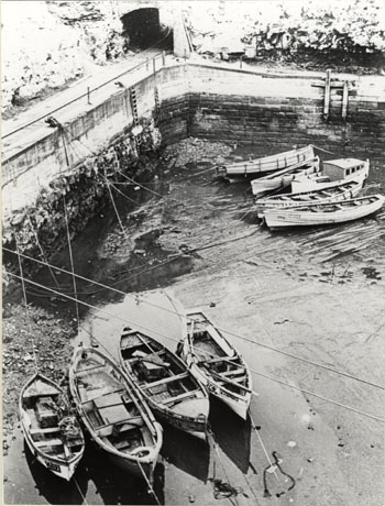 Photograph showing the walls of a dock, with behind them the mouth of a tunnel; in front of them seven small boats are resting on the surface of the dock at low tide; the dock has been identified as Inner Dock with Railway Tunnel, Seaham