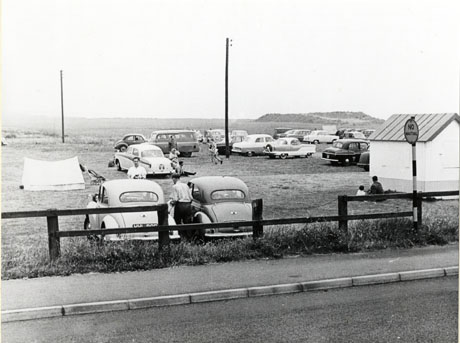 Photograph of a field at the top of a cliff next to the sea, with approximately sixteen cars parked in it; eight people are sitting and walking near the cars and a hut is on the right of the picture; the surface of a road and a broken fence are at the front of the picture, which has been identified as The Hall Car Park, Seaham, 1960s