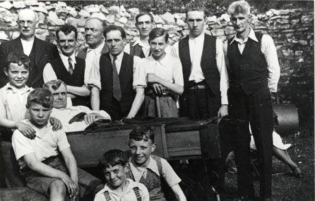 Photograph of eight men and five boys in front of stone wall; one boy is holding an implement of some kind and aman is in an open carriage which resembles a cart; most of the men are in shirt sleeves; the photograph has been described as Rock House Garden Party Seaham (Frank Sonny Anderson in the Wheel Carriage), 1930s