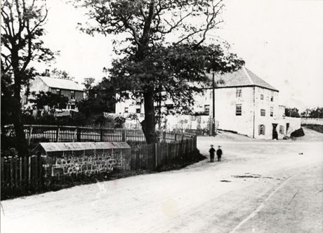 Photograph showing the surface of a road with a wall, fence and two trees in the foreground; beyond the trees is a large plain oblong building with a sign board on its front; behind that building a smaller house can be seen; the photograph has been described as Mill Inn, Seaham, early 1900s