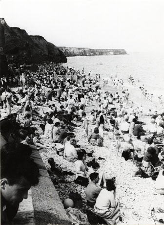 Photograph of a strip of beach with hills behind on the left of the photograph and the sea on the right; the beach is crowded with people with their backs to the camera looking out to sea; the photograph has been identified as Seaham Beach, Bank Holiday, 1961