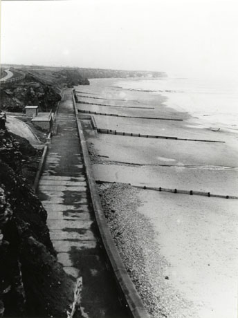 Photograph, taken from above, of a promenade stretching away from the camera with the hills on the left, and the beach and the sea on the right, of the photograph; breakwaters can be seen on the beach; the photograph has been identified as looking towards Ryhope