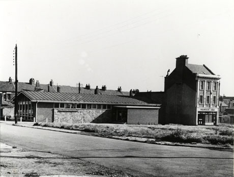 Photograph, taken from across the road, of the glass front and stone and brick side of a low building, with the words Seaham Branch Library on its side; at the end of the library is a building of three storeys with the name Volunteer Arms on its front; between the two buildings and the camera is open space, possibly the result of demolition; house roofs can be seen in the distance behind the library