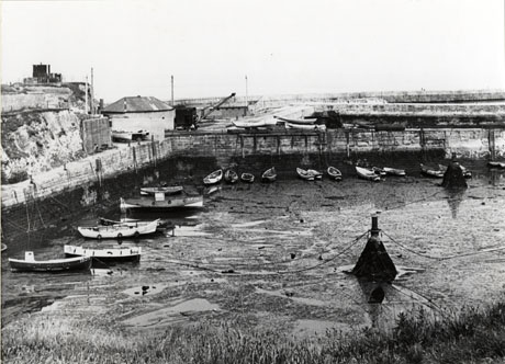 Photograph of the walls of a dock and, behind them, a view of the top of the dock walls, the slope leading down to the dock and a building on the dockside; on the surface of the dock nineteen boats can be seen because of low tide; the photograph has been identified as the Inner Dock at Seaham, but is looking out to sea in a different direction from the photograph numbered seah0155