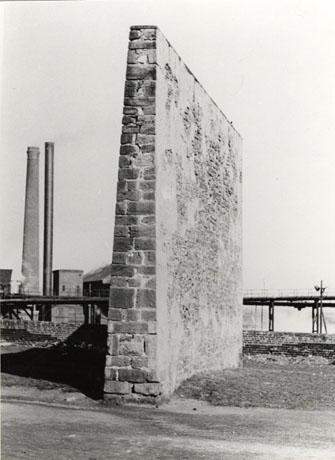 Photograph of the end of a wedge shaped high wall standing on its own with two tall chimneys and horizontal pipes in the background; the wall is made of stone and is roughly rendered; it has been identified as Hand Ball Alley Near Seaham Pit Yard, Demolished 1953