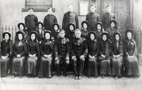 Photograph of a group of sixteen women and seven men in Salvation Army uniform, outside a building with a large door and a railing; the group has been identified as being in Seaham