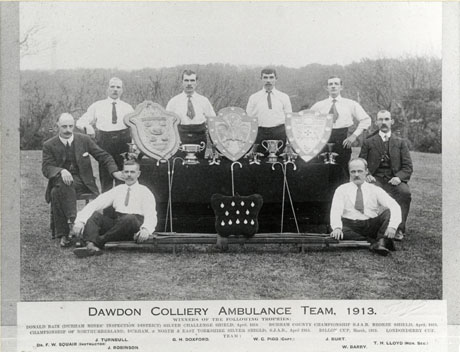 Photograph of ten men standing and sitting in the open air round a table on which two trophy cups, five trophy candlesticks, three shields and twelve medals are displayed; two men in front of the table are sitting near a rolled up stretcher; a caption on the picture identifies the men as Dawdon Colliery Ambulance Team, 1913, winners of the Donald Bain (Durham Miners Inspection District) Silver Challenge Shield, April 1913; Durham County Championship S. J. A. B. Bronze shield, April 1913; Championship of Northumberland, Durham and North and East Yorkshire Silver Shield S.J.A.B., April 1913; Dillon Cup, March 1913; Londonderry Cup; the members of the team are as follows: J. Turnbull; G. H. Doxford; W. C. Pigg (Captain); J. Burt; Dr. F. W. Squair (Instructor); J. Robinson: W. Barry; T. H. LLoyd (Honorary Secretary)