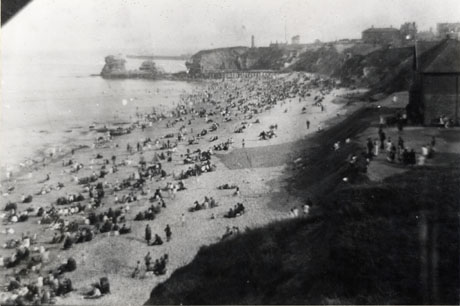 Photograph of a beach with the cliffs on the right of the picture and the sea on the left; the cliffs curve round the bay to the left and detached rocks are in the sea at the end of the curve of the bay; people and buildings can be seen on the right of the picture on the top of the cliffs; the beach is covered with crowds of people looking out to sea; the beach has been identified as Seaham Beach