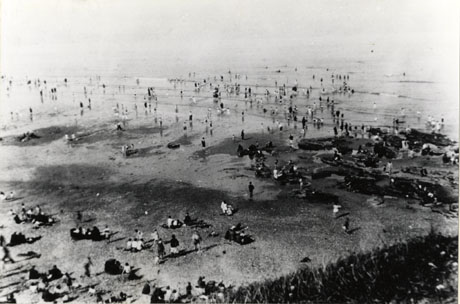 Photograph of the surface of a beach, rocks on the beach and the sea; people are on the beach and in the water; the photograph is taken from above and behind the scene and all the people are looking away from the camera; the beach is identified as Seaham Beach