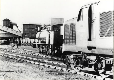 Photograph showing the side of a steam locomotive standing on a rail track near a colliery; on a parallel track nearer to the camera is the side of a diesel locomotive; part of the colliery buildings can be seen in the distance; the locomotives have been identified as Gamma and a diesel at Vane Tempest Colliery, Seaham, in 1974; a man is working on the top of Gamma; rows of coal waggons can be seen just beyond Gamma