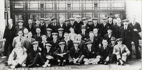 Photograph of a group of forty three men in suits, ties and caps posed in a building in front of a glass partition; they are identified as Seaham miners at the Convalescent Home at Saltburn
