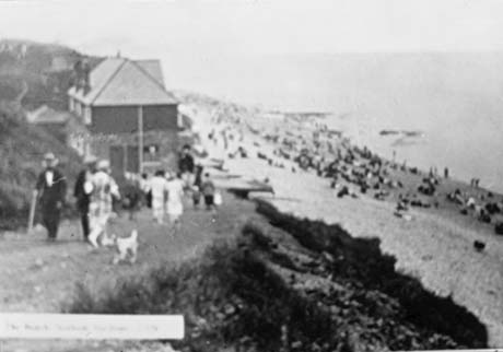 Postcard photograph with an illegible caption, showing a blurred picture of crowds on the beach in the distance at the right of the photograph, a house at the top of the cliff and approximately twelve people walking up and down the cliff path in the foreground at the right; the photograph has been described as showing the beach at Seaham