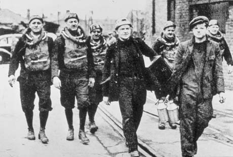 Photograph showing five men in mining rescue gear walking along past a brick building with a man carrying a box and a boy of approximately fifteen years, carrying two metal drink containers; the side of a car and indistinct people can be seen behind them; they have been identified as Dawdon Miners