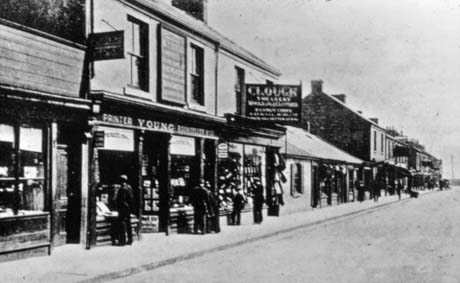Photograph of one side of a street with shops and houses on it; there are three shops close to the camera, one of which cannot be identified; one is kept by Young, a Printer and Bookseller, and the other by Clough, the occupation of whom cannot be deciphered; the shops further along the street cannot be determined; there are figures on the pavements; the photograph has been described as North Railway Street, Seaham