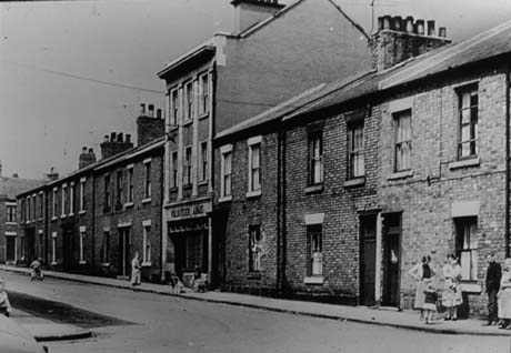 Photograph of one side of a street of brick terraced houses with a taller building in the middle of the terrace with pilasters on its front and the words Volunteer Arms above its entrance; there are four women, one man and six children on the pavement; the street has been identified as Francis Street, Seaham, before Demolition