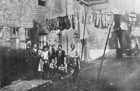 Photograph showing the facades of a square of brick buildings in poor condition, some of which have broken windows; a group of six women are standing near the buildings with three small children, aged approximately between two and four years; behind the group is a line of washing; the photograph has been identified as depicting Summersons Buildings, off Railway Street, Seaham
