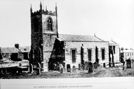 Photograph of the exterior of the west end and south side of St. John's Church, Seaham Harbour, showing the crenellated tower and part of the graveyard; behind the church tower, terraced houses can be seen and beyond the east end larger terraced houses can be seen
