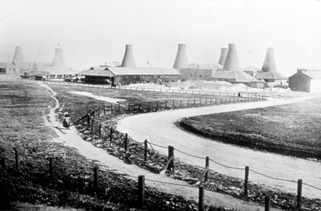 Photograph showing a road running round to the right leading to low buildings with seven bottle-shaped kilns; a pile of some substance can be seen in front of the buildings; they have been identified as Candish's Londonderry Bottleworks, which were built in 1853 and demolished in 1921