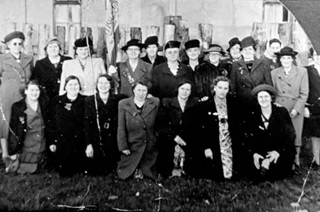 Photograph of twenty women, in hats and overcoats, posed in two rows in front of a fence with a house behind; one woman is holding a furled flag; they have been identified as members of the Women's Branch of the Royal British Legion; the flag appears to read Woodham Branch, Newton [Aycliffe]
