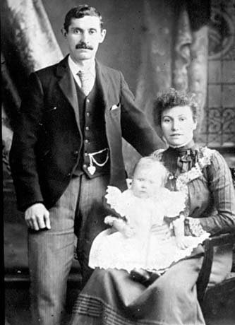Photograph showing a man wearing a dark jacket and waistcoat and pinstripe trousers, standing on the left of the picture with a woman, wearing a dress with tucks and lace, sitting on a wooden chair with arms; she is holding an infant wearing a frilly dress; the child has been identified as Mr. Masters as a Baby