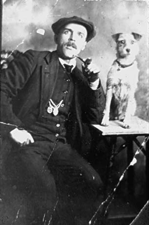 Photograph of a man wearing a dark suit and waistcoat and cap, sitting with his left arm resting on a small table on which a small terrier dog is sitting; the man is smoking a pipe and has a moustache; he has been identified as Tim Dowding of Seaham