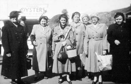 Photograph showing seven women in overcoats standing with an indistinct background behind them; an advertisement for O' Hagan's Oysters can be seen behind them; one woman is holding a sunshade and another two carrier bags; they have been identified as Miners' Wives and Widows Trip, Seaham
