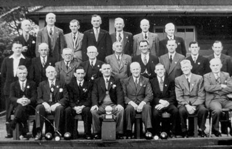 Photograph showing twenty five men in suits posed in three rows in front of a single-storey building and on the edge of the bowling green; there are four bowls , two jacks and a trophy, in front of the group, which has been identified as members of Dawdon Bowling Club