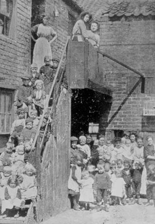 Photograph showing the corner of a square of brick houses with a staircase running up the front of the house on the left; a crowd of children, aged approximately between three and six years, are standing on the steps; three women are standing at the top of the steps; on the ground beneath the steps, another crowd of children of the same ages are standing with approximately seven women; they have been identified as being in Hawkey's Yard, North Railway Street, Seaham