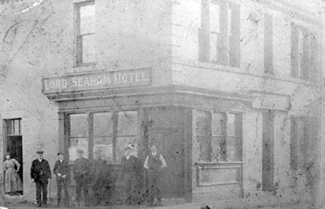 Photograph showing the front and side of a building with large sash windows, with an entrance at the front and a board saying: Lord Seaham Hotel at the side; beneath the notice, six men are standing against the wall, and a woman is standing in a doorway in the side of the building; the hotel has been identified as being in Seaham