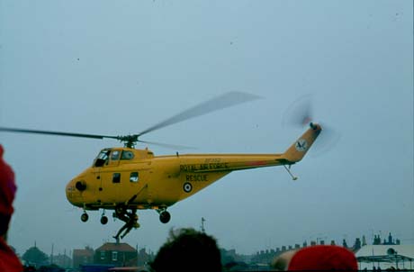 Photograph showing a Royal Air Force Rescue helicopter, no. XP 352, hovering while a man is being winched up; the tops of houses in the distance and the tops of the heads of three people in the foreground can be seen; the venue of the helicopter's activity has been identified as Seaham Show