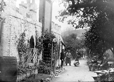 Photograph of the front of a brick building with crenellations, trellises, creepers, and a board, on the left of the picture; in front of the building is a path on which two men in leather aprons are standing, with another man and a barrel lying on the ground; two other men in suits are sitting on a bench under a tree and two girls are at the end of the path behind the men in aprons; the photograph has been identified as Peartree Public House and Adam and Eve Gardens, Seaham; the man standing with the men in aprons has been identified as Ralph Fair, Manager