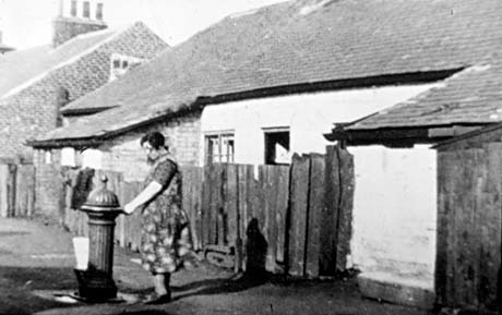Photograph showing, on the right, the back of a low house with two jutting lean-tos; a window and an open door can be seen;in the centre of the picture a woman wearing a pinafore is standing at a water pump filling a container