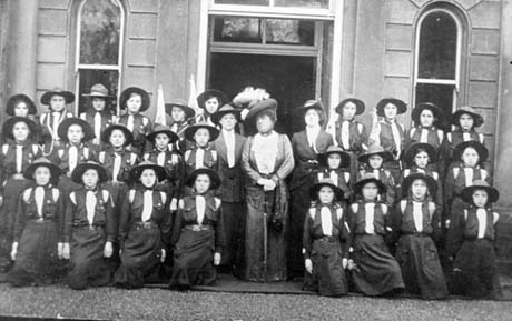 Photograph showing twenty six young women, wearing uniform, posed in three rows in front of a large house with narrow round windows either side of a large door; a middle-aged woman wearing a large hat and long dress is standing in the middle of the group with two women in uniform on either side of her; they have been described as First Girl Guides, Seaham