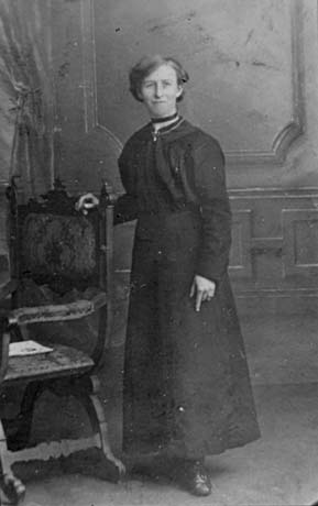 Photograph, full-length, of a woman wearing an ankle-length, high-necked, long-sleeved, dark dress; she is also wearing boots, a necklace, and a ribbon round her neck; she is standing resting her arm on the back of a chair in a photographer's studio; she has been identified as Mrs. Phoebe Hudson
