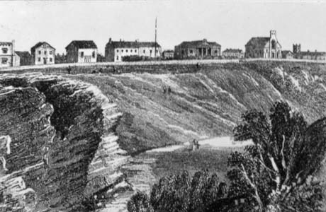 Photograph of an engraving of the side of a deep slope with trees in the foreground and ten buildings at the top of the slope; they include two churches and a building with classical columns; it has been identified as Seaham Harbour from the Dene