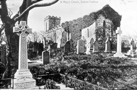 Postcard photograph entitled St. Mary's Church, Seaham Harbour, showing the exterior of the east end and south side of the church covered in a creeper; part of the grave yard can also be seen, with a celtic cross in the foreground on the grave of Charles Stewart Reginald Stewart