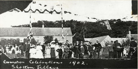 Photograph showing groups of men, women and children, possibly on a green, in the open air in front of a row of cottages, with other cottages at the right of the picture, and trees behind the buildings; above the people are bunting on poles and Union Jacks; under the picture is a hand-written caption, as follows: Coronation Celebrations,1902, Shotton Village; the Coronation was that of King Edward VII