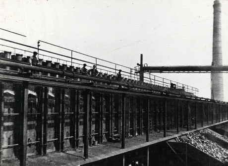 Photograph of a row steel uprights with masonry behind them and a platform in front of them; a shute containing coal or coke coming from the platform can be seen to the right of the picture; four men are standing on the top of the structure, which has been identified as Kopper's Coke Ovens, Shotton Colliery; a tall chimney stands at the end of the structure