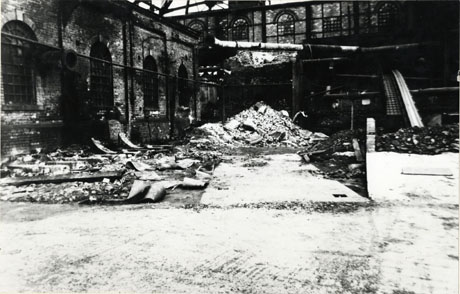 Photograph of the exterior of the wall of a large brick building with four large round- headed windows, on the left of the picture; this building is at right angles to another higher wall, also containing round-headed windows, at the back of the picture; in the space between the walls is debris of old pipes, bricks, planks of wood and piles of similar rubbish; the picture has been described as Looking North To Heapstead In North Pit Headgear Shotton, 12 April 1957