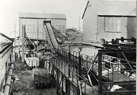 Photograph showing on the left of the picture the side of a brick building with a pipe along its length; at the end of the building at the back of the picture is a cabin raised up; a shute is coming from the top of the cabin down to a raised platform with steel fencing round it which goes under the cabin; another cabin made of corrugated iron is at the right of the picture; the picture has been described as Coke Cutting and Screening Plant, Shotton, 1928