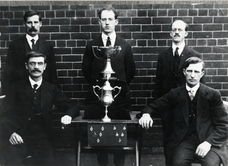 Photograph of six men in suits and ties, posed in front of a wall, with a table, on which there is a trophy cup, identified as the Dr. Irvine Cup, another cup, and ten medals; the men are identified as Shotton Colliery Ambulance Team