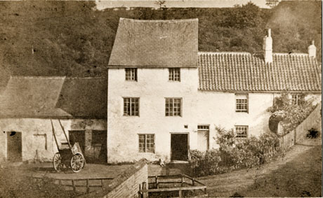 Photograph of the exterior of a building, with a slope covered with trees behind it, and a slope rising in front of it, to the right of the picture; to the left of the house is a low building of rough masonry, most likely a stable; the middle portion of the house has three storeys with two windows on each of the two upper storeys; to the right is a lower wing of two storeys, partly obscured by the rising road in front of it; a cart is parked in front of the stable; the house has been identified as Shotton Mill