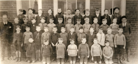 Photograph of forty three boys, aged approximately ten years, posed in front of a brick building with a man, presumably their teacher; a boy on the front row is holding a notice which reads Shotton Boys' Council School Class 6
