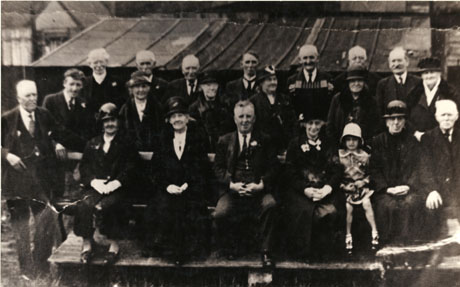 Photograph of a group eleven middle aged men and nine middle aged women and one child sitting on benches with the roof of a tent in the background; they are all dressed formally in coats, hats and suits; a man on the back row is holding an accordion; they are described as Shotton Methodist Group