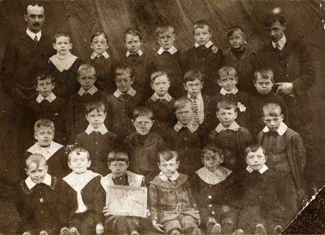 Photograph of twenty five boys, aged approximately seven years, posed in front of fabric, and accompanied by two men, presumably their teachers; they have been identified as pupils at St. Saviour's School, Shotton, which has been further identified as the Tin School, now the Comrades' Club