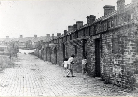 Photograph showing the cobbled surface of a lane with the back walls the yards of terraced houses on the right hand side and vegetation on the left hand side; a row of houses is in the distance and the tops of the houses behind the wall can be seen; three children are playing in the lane, but are indistinct