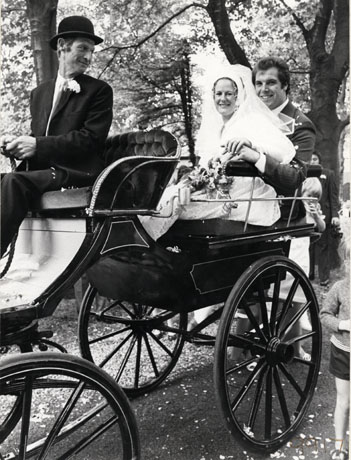 Photograph of a woman in a wedding dress and veil, sitting in a horse drawn trap with her husband behind her; a man in a suit, bowler hat and button hole is sitting in the driving seat of the trap; parts of two children can be seen near the rear of the trap; the bride and her husband have been identified as Judith Howe and Alan Briggs and their wedding as having taken place at the church of St. Saviour, Shotton