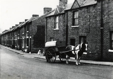 Photograph showing terraced houses along a street on the right of the photograph ; a horse-drawn covered two-wheeled cart is standing in front of a house near the front of the photograph; three indistinct small children are on the pavement; the street has been identified as Dene Street, Shotton, and the cart as belonging to the Sherburn Hill Co-operative Society